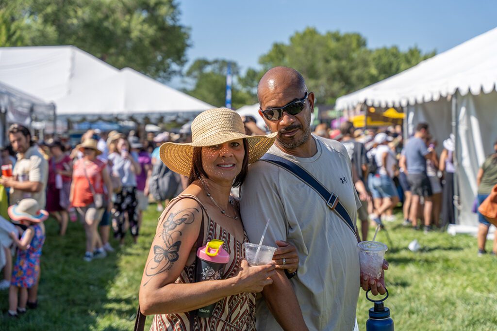 Mixed-race woman with a tattoo on her arm smiling wearing a white hat holding a drink and Black man in a light blue shirt and sunglasses standing close to her. hey are outside in the sunshine and in the background there are white tents with crowds of people.